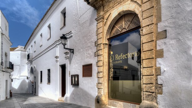 A narrow street with a building featuring a large arched window with the text "El Refectorio Restaurante". - TUGASA Convento San Francisco (Vejer de la Frontera)