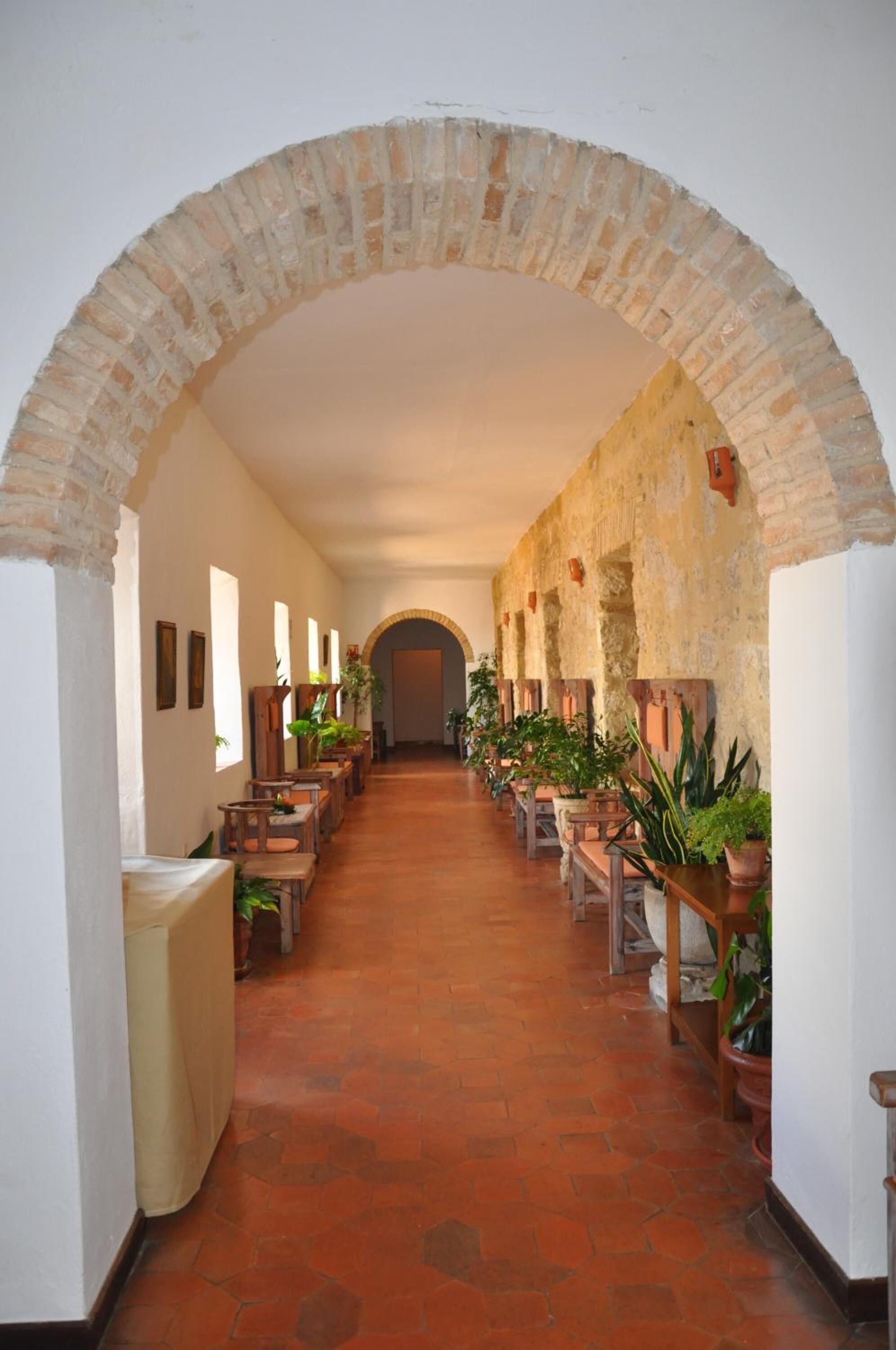 A corridor with a brick archway, wooden benches, potted plants, and a tiled floor.