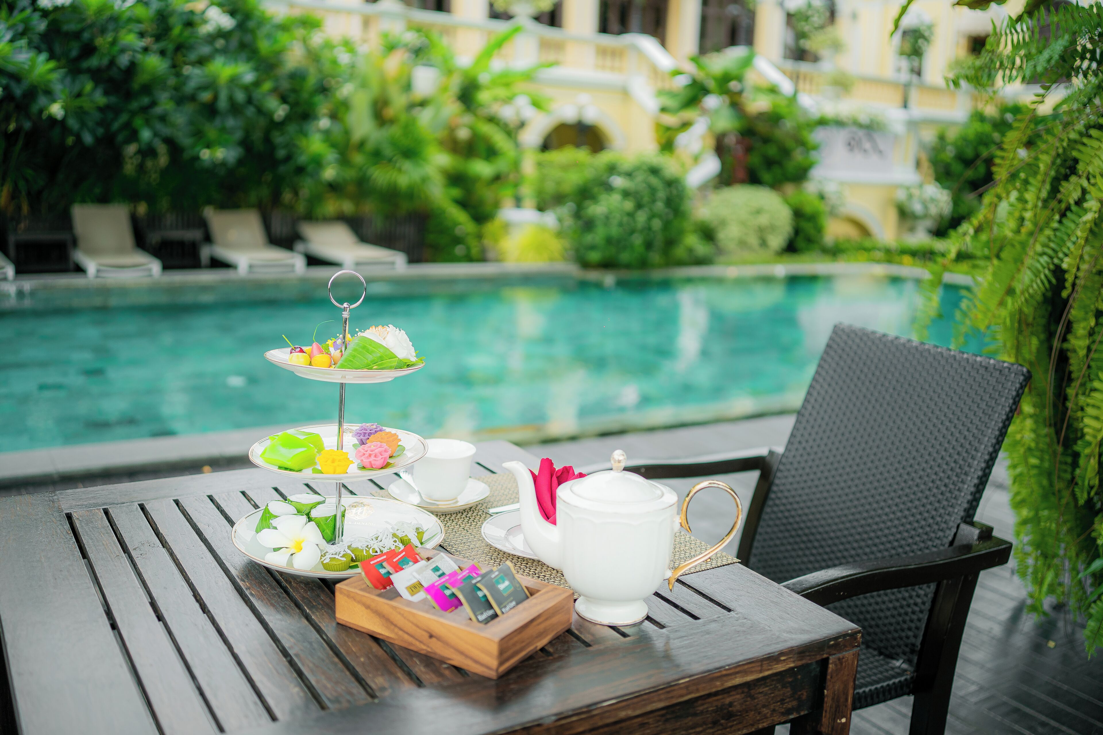 A table with a teapot, cups, a tiered tray with snacks, and a box of tea bags by a poolside with lounge chairs and greenery in the background.