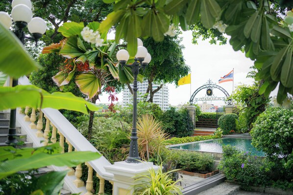 A lush garden with a fountain, a staircase, and a building with flags in the background.