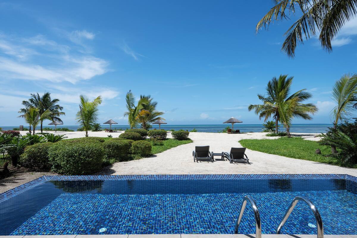 Ein Pool mit Liegestühlen und Sonnenschirmen, umgeben von Palmen und Blick auf den Ozean.