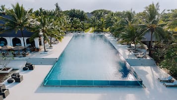 Uma piscina comprida com espreguiçadeiras e guarda-sóis em ambos os lados, cercada por palmeiras e um edifício com arcos.