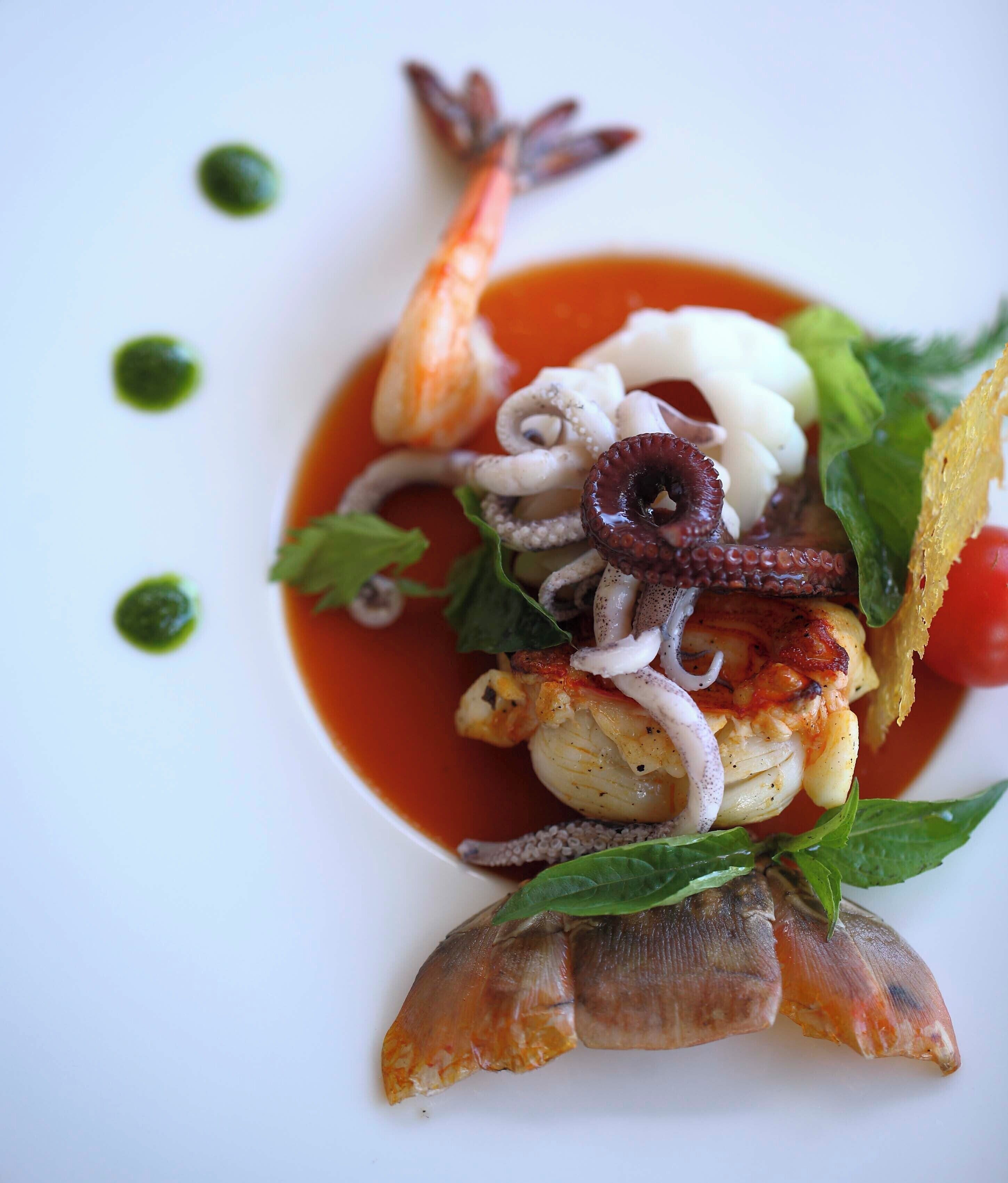 A seafood dish with shrimp, octopus, and a fish head, garnished with basil and served with a red sauce.