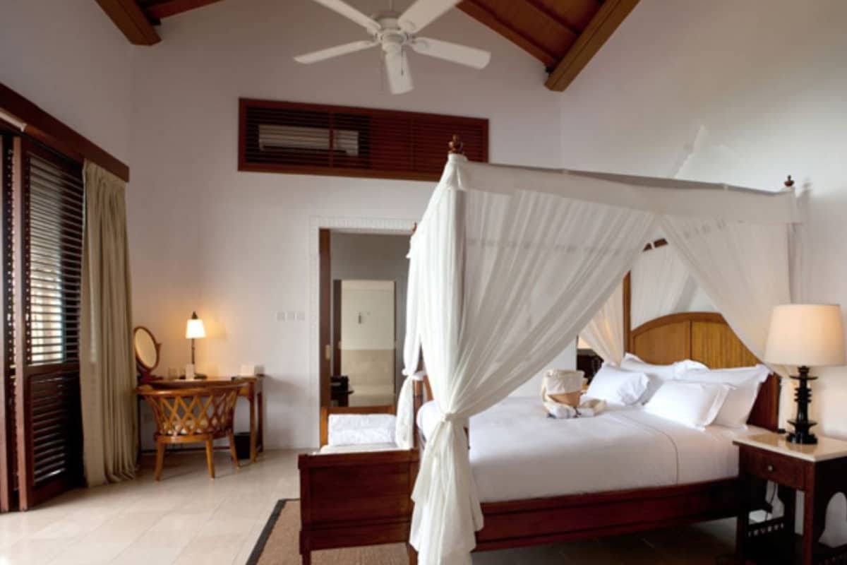 Un dormitorio con cama con dosel, ventilador de techo, mesita de noche con lámpara y una silla de madera.