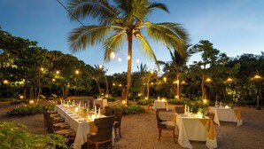 Un espace de restauration en plein air, avec des tables pour deux, entouré de verdure et de palmiers, et illuminé par des guirlandes lumineuses.