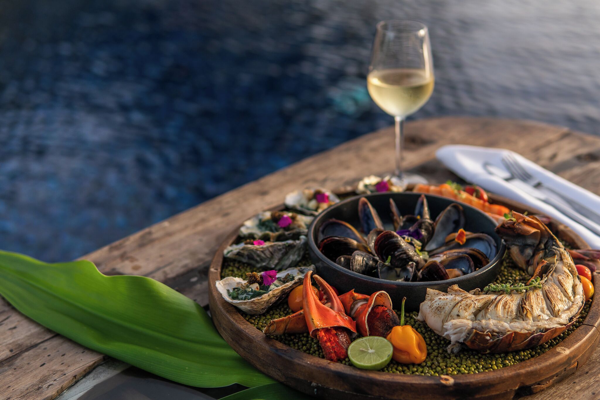 A seafood platter with mussels, oysters, and a fish on a wooden board, accompanied by a glass of white wine, set against a blurred background.
