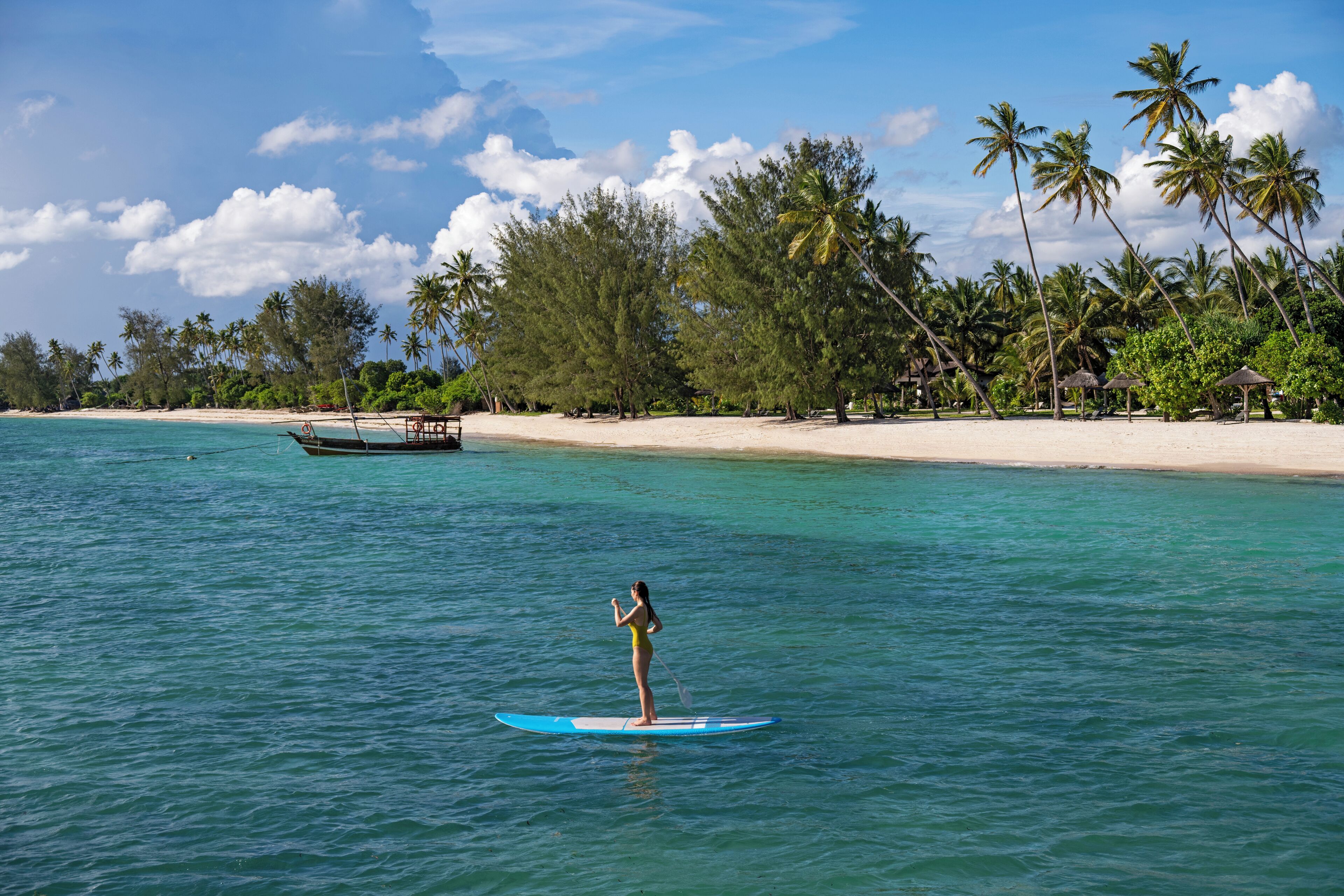 A beach with clear turquoise water, a woman paddleboarding, and palm trees in the background.