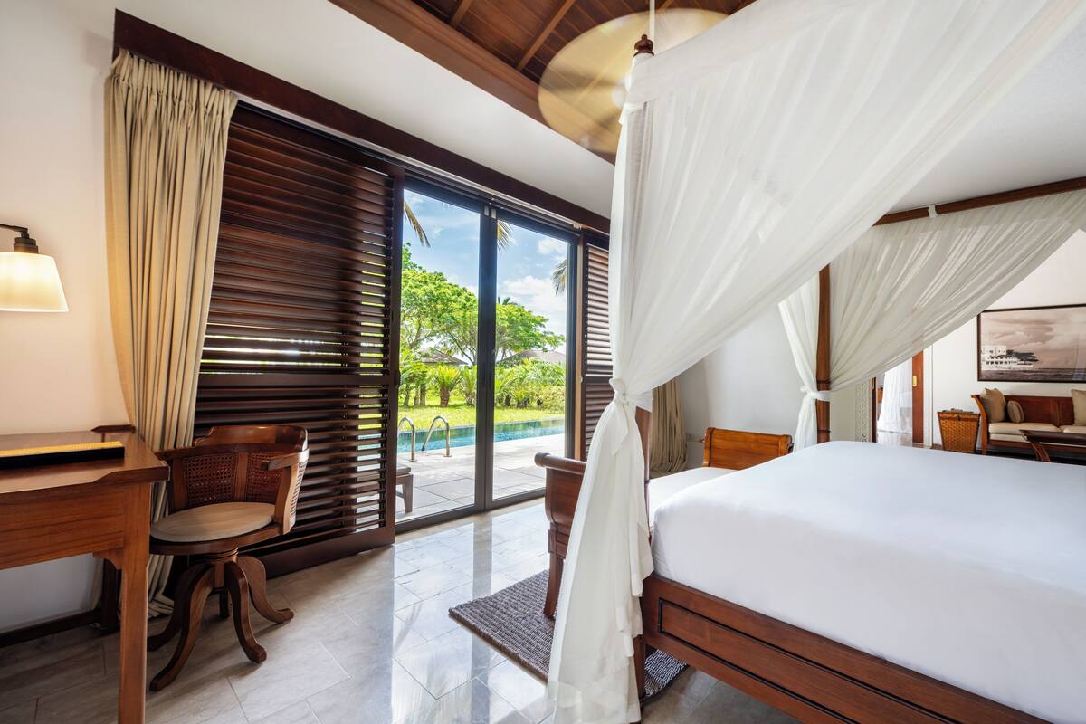 Ein Schlafzimmer mit einem großen Bett, einem Holztisch, einem Stuhl und Blick auf ein Schwimmbecken und Grünflächen durch das Fenster.