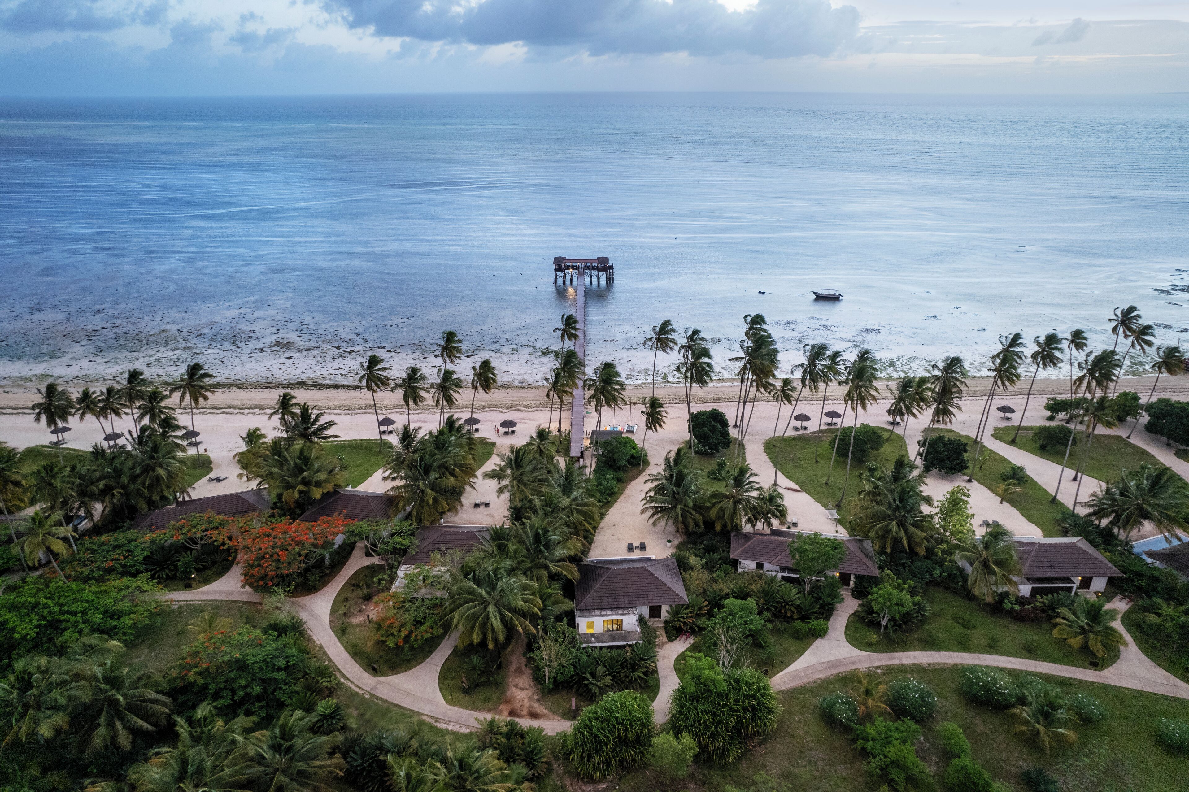 An aerial view of a resort with a beach, palm trees, and a pier extending into the sea.