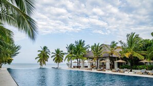 Un espace aménagé au bord de la piscine, avec des chaises longues et des parasols, des palmiers et un bâtiment doté d’un toit en tuiles.