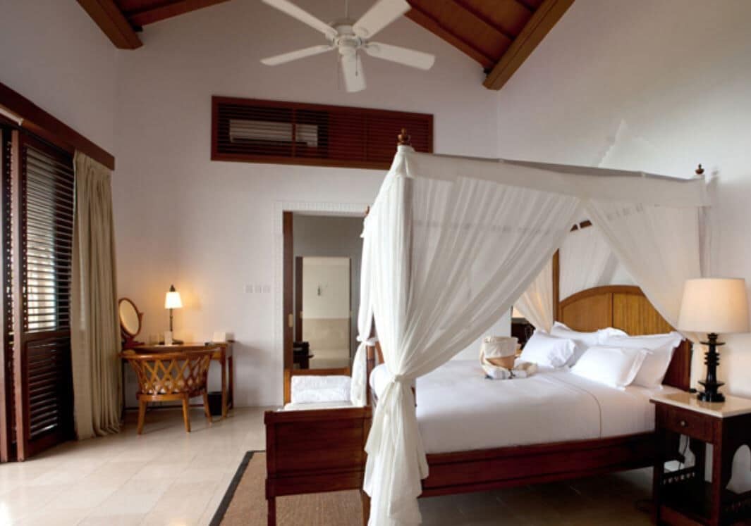 A bedroom with a four-poster bed, a ceiling fan, a bedside table with a lamp, and a wooden chair.