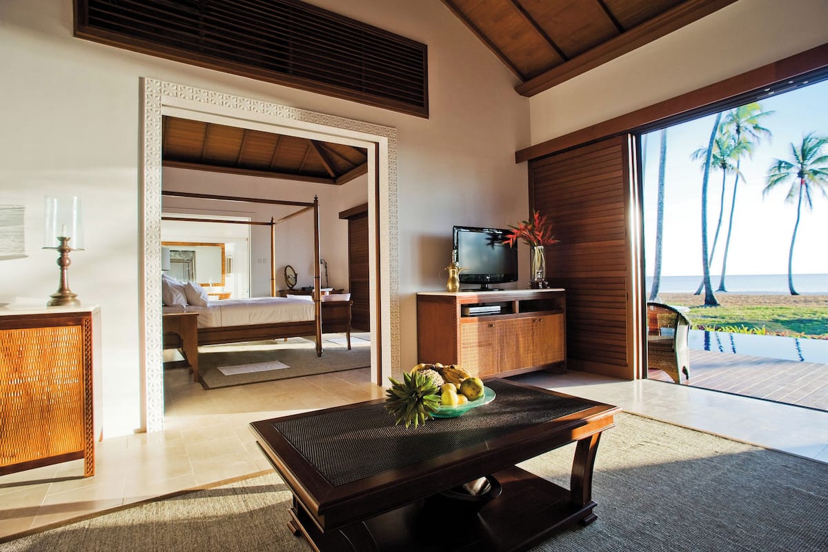 Ein Zimmer mit einem Bett, einem Fernseher, einem Esstisch mit Obst und Blick auf den Strand.