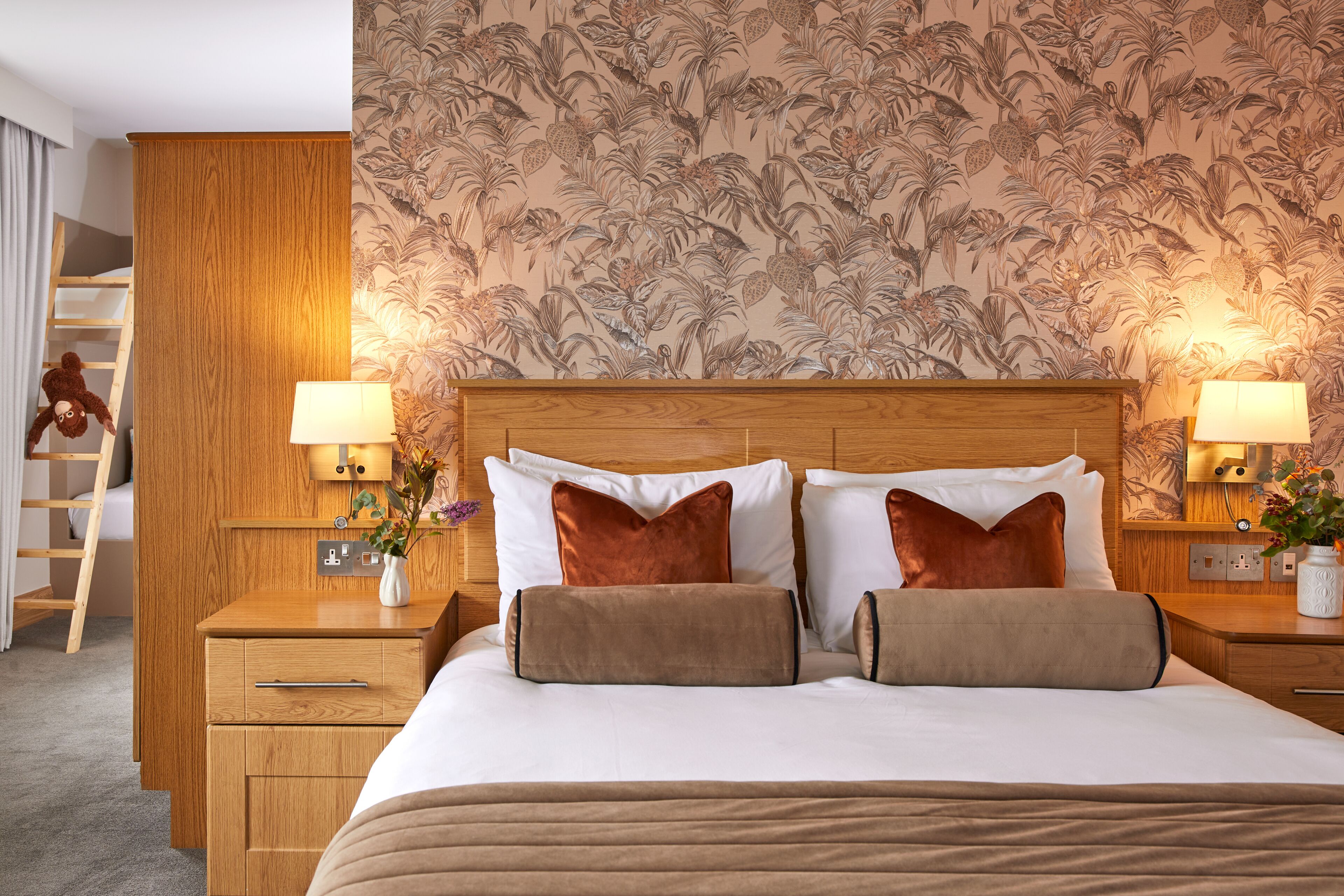 Habitación de hotel con cabecera de madera, dos mesitas de noche con lámparas, cama con ropa blanca y almohadas marrones, armario de madera y papel tapiz floral.