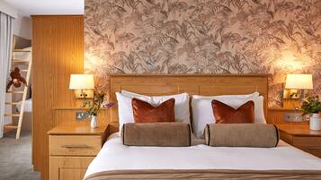 Habitación de hotel con cabecera de madera, dos mesitas de noche con lámparas, cama con ropa blanca y almohadas marrones, armario de madera y papel tapiz floral.