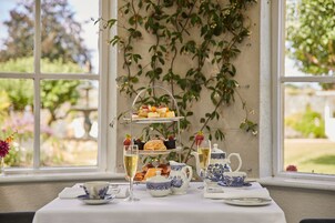 Un expositor de tres niveles con pasteles y frutas, una tetera, tazas y una jarrón sobre una mesa junto a una ventana con vistas a zonas verdes.