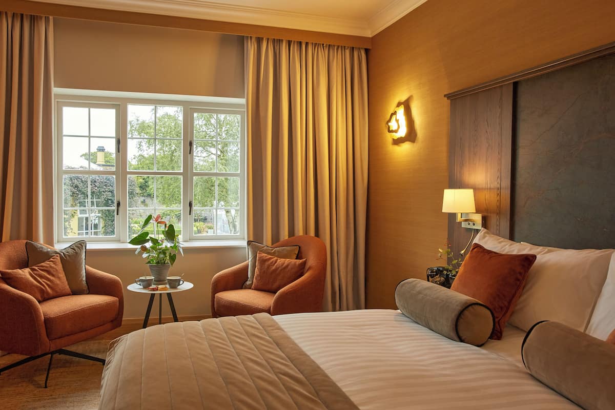 A hotel room with a large bed, two armchairs, a small table with a plant, and a window with curtains.