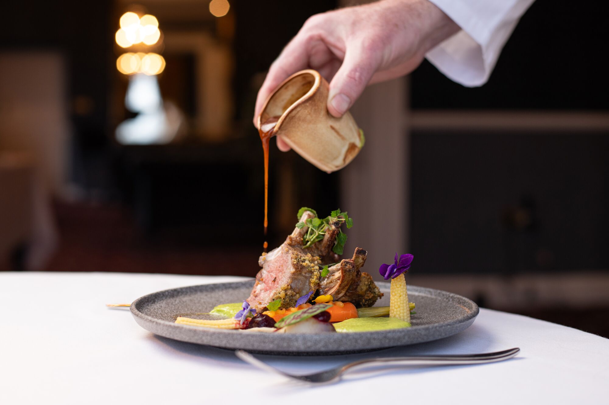 A chef pouring sauce over a dish with meat and vegetables on a plate.