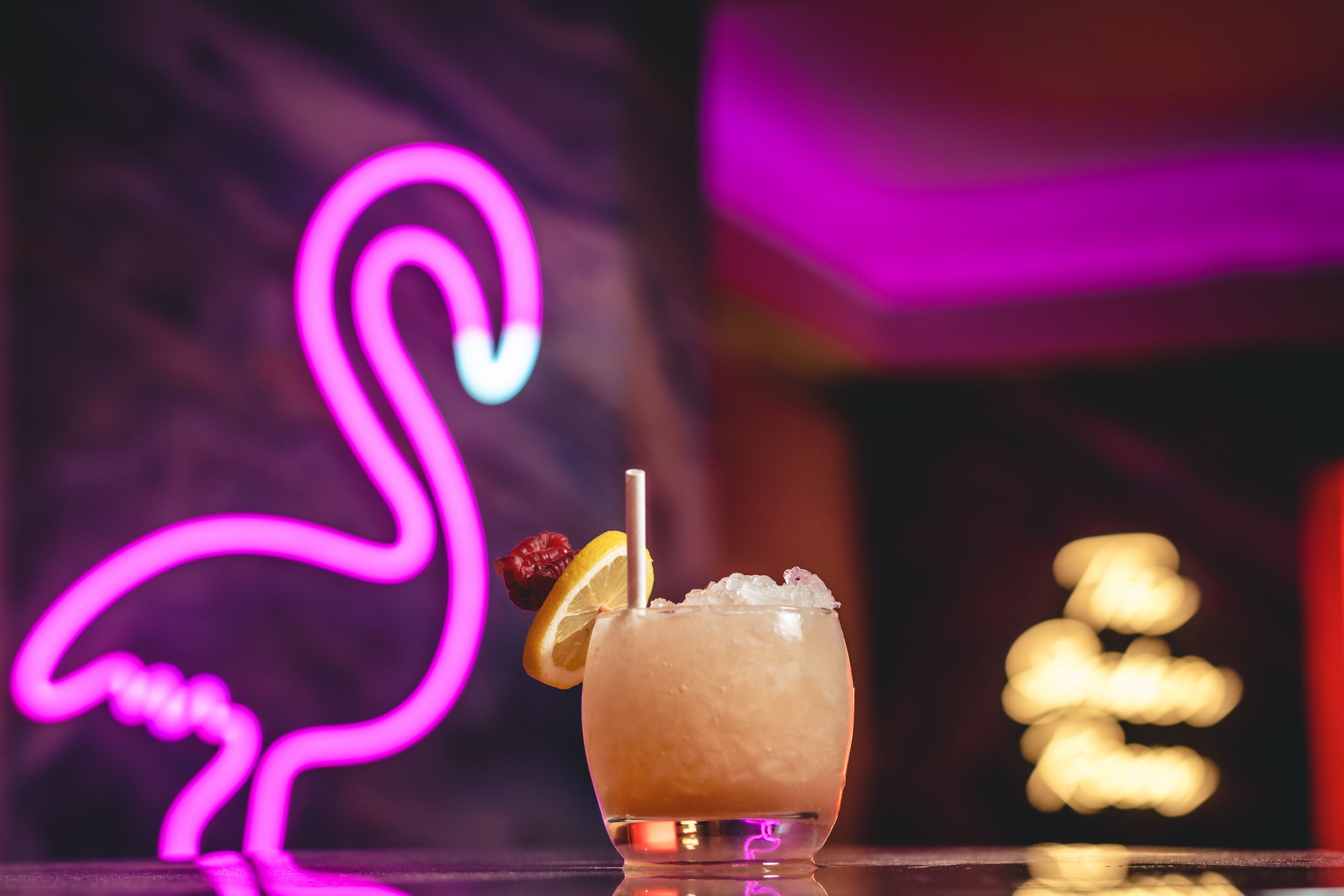 A cocktail with a straw and a slice of lemon on the rim, ice cubes, and a cherry garnish, set against a background with a neon flamingo sign.