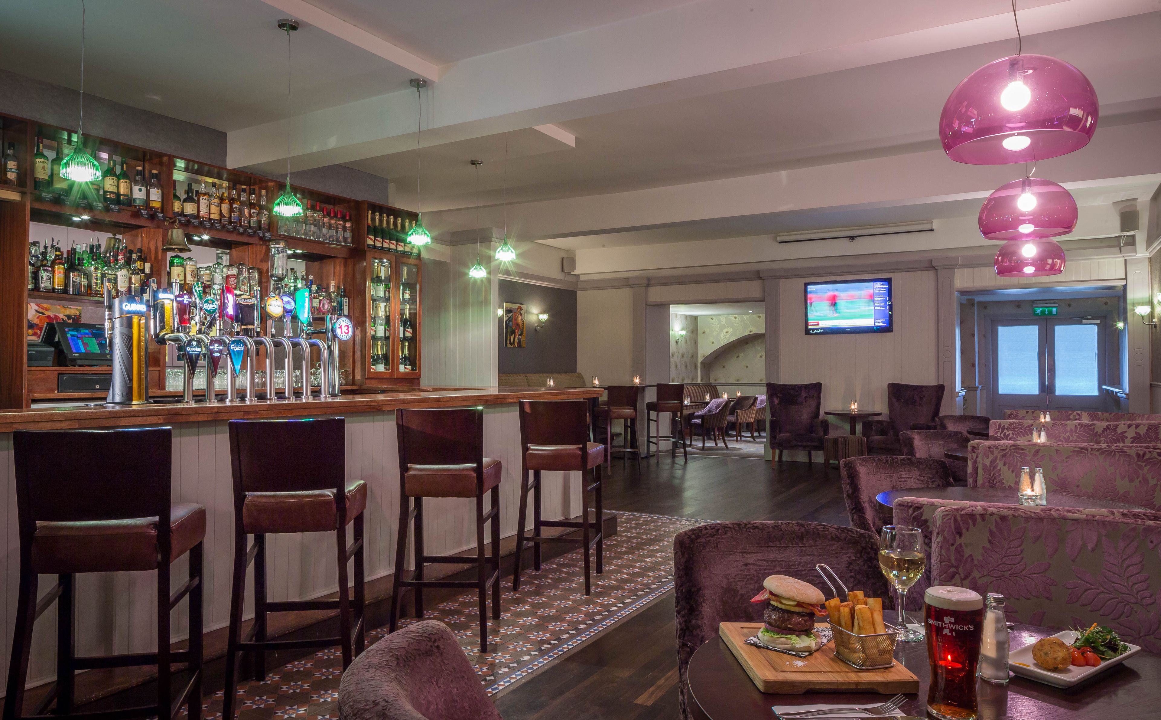 A bar area with a variety of liquor bottles and a digital screen displaying a sports game. There are multiple seating areas with bar stools and cushioned chairs.
