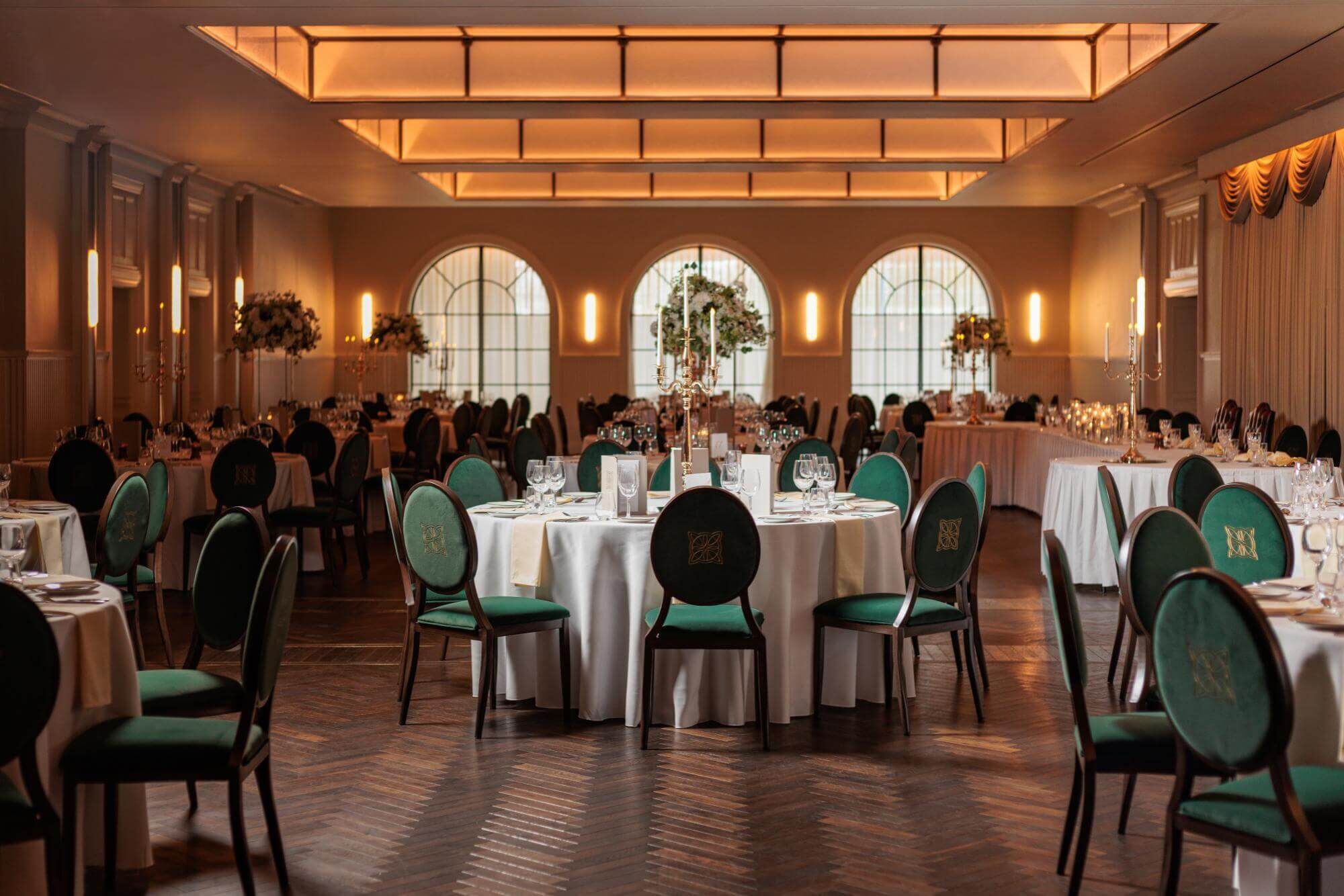 A banquet hall with round tables, green upholstered chairs, and chandeliers.