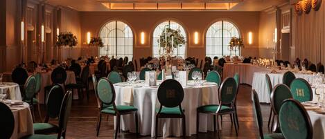 A banquet hall with round tables, green upholstered chairs, and chandeliers.
