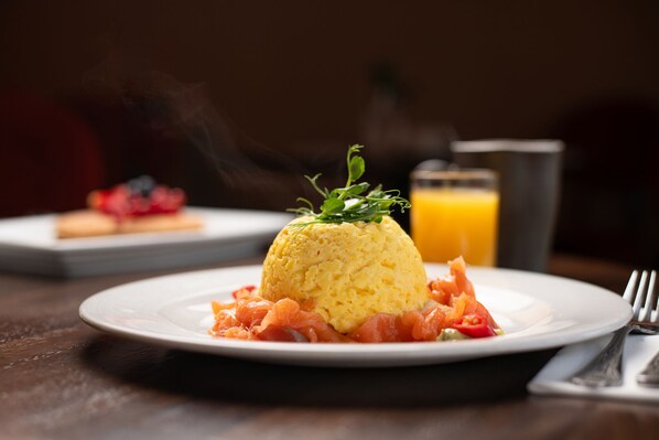 A plate with a mound of yellow food, possibly scrambled eggs, garnished with green herbs, accompanied by slices of salmon and red pepper. In the background, there's a blurred glass of orange juice and a dessert plate. - County Arms Birr (Birr)