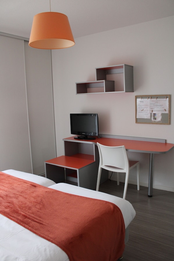 A hotel room with a bed, a desk with a computer, a chair, and a bulletin board on the wall.
