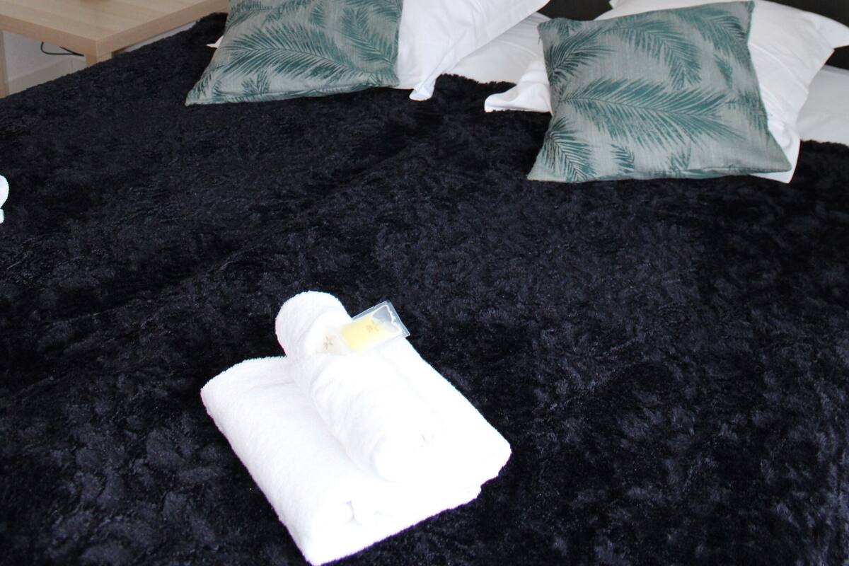 Een slaapkamer met een donker hoofdeinde, twee witte kussens met groene palmbladmotieven, een zwart geweven beddeken, een witte handdoek en een bedlampje.