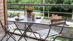 Un balcón con una mesa dispuesta para dos personas, destacando un jarrón de flores, un plato de pasteles y dos tazas.