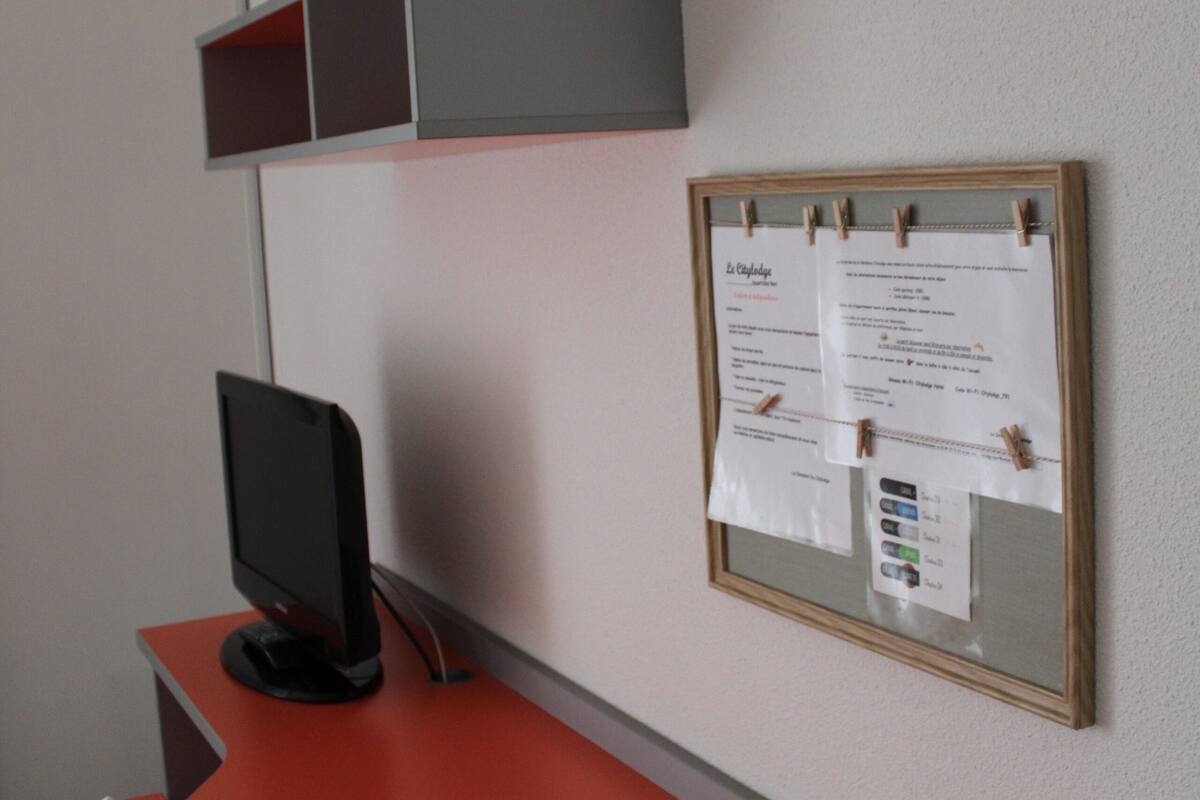 Een werkplek met een rode bureau, een beeldscherm en een prikbord met eraan geklemde papieren.