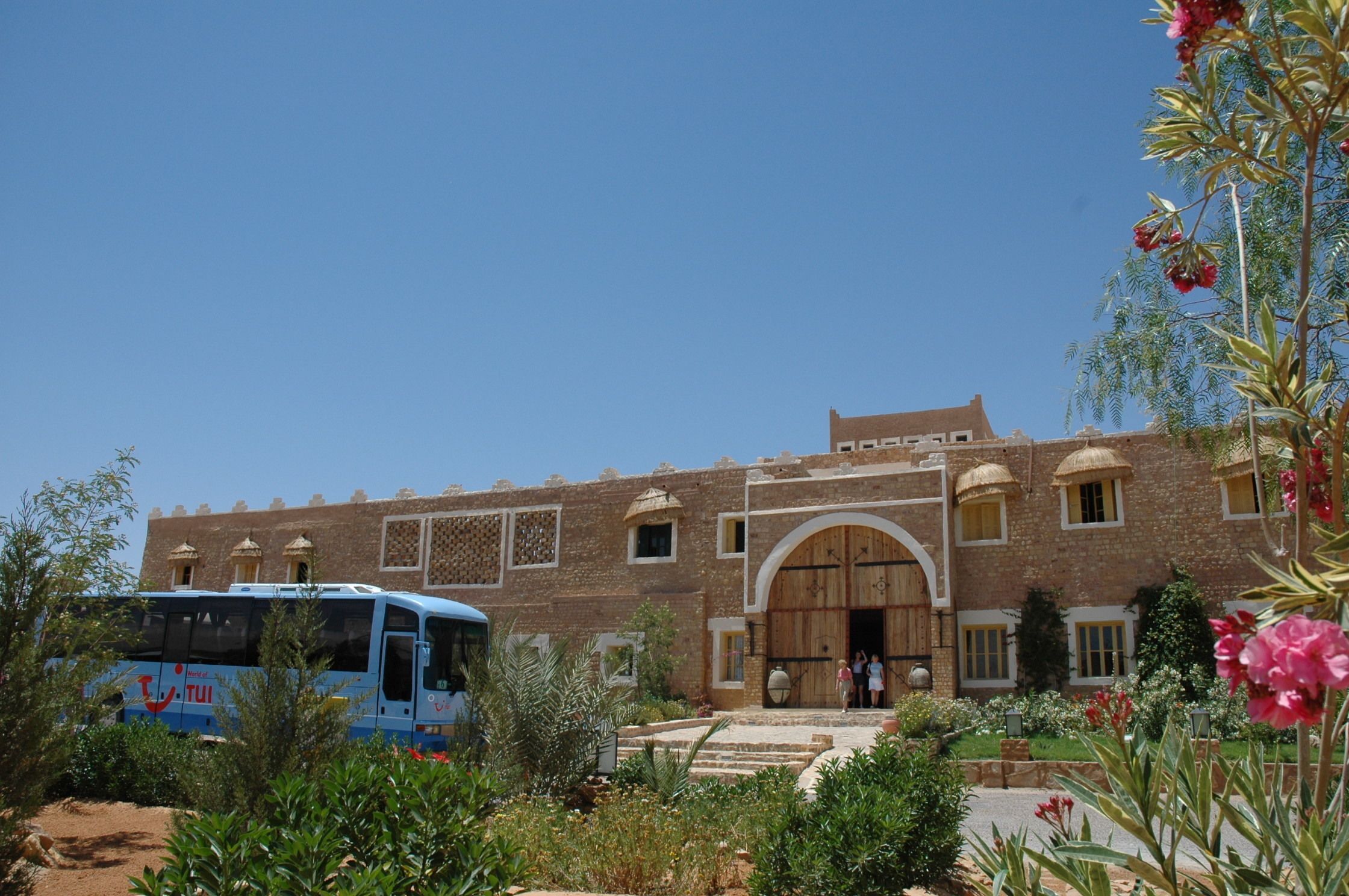 Un bâtiment traditionnel doté d’une grande porte d’entrée en bois, avec un autobus garé devant, et un jardin où poussent diverses plantes et fleurs.