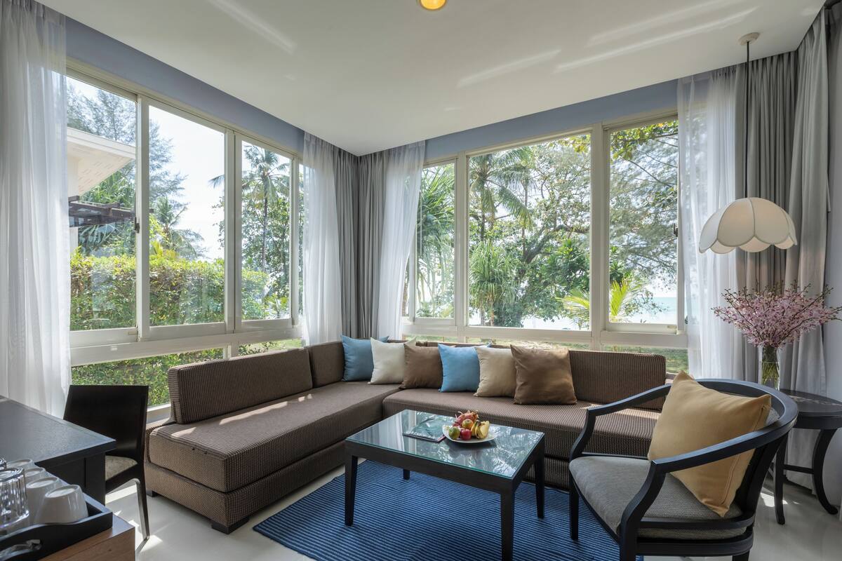A modern living room with a sectional sofa, a glass coffee table, and a dining area with a table and chairs. Large windows provide a view of greenery outside.