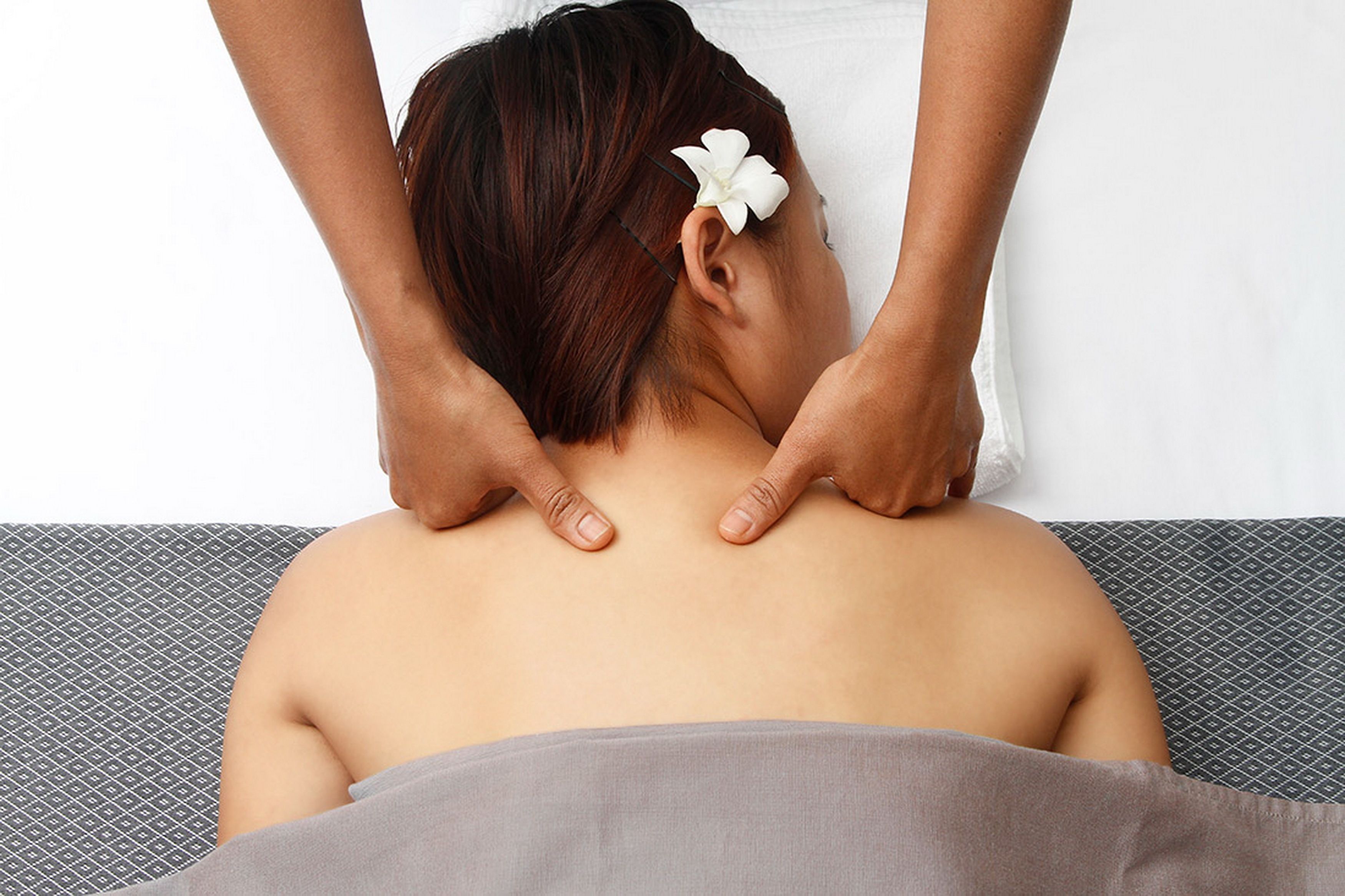 A person receiving a back massage with a white flower accessory in their hair, lying on a bed with a patterned headboard and a gray blanket.