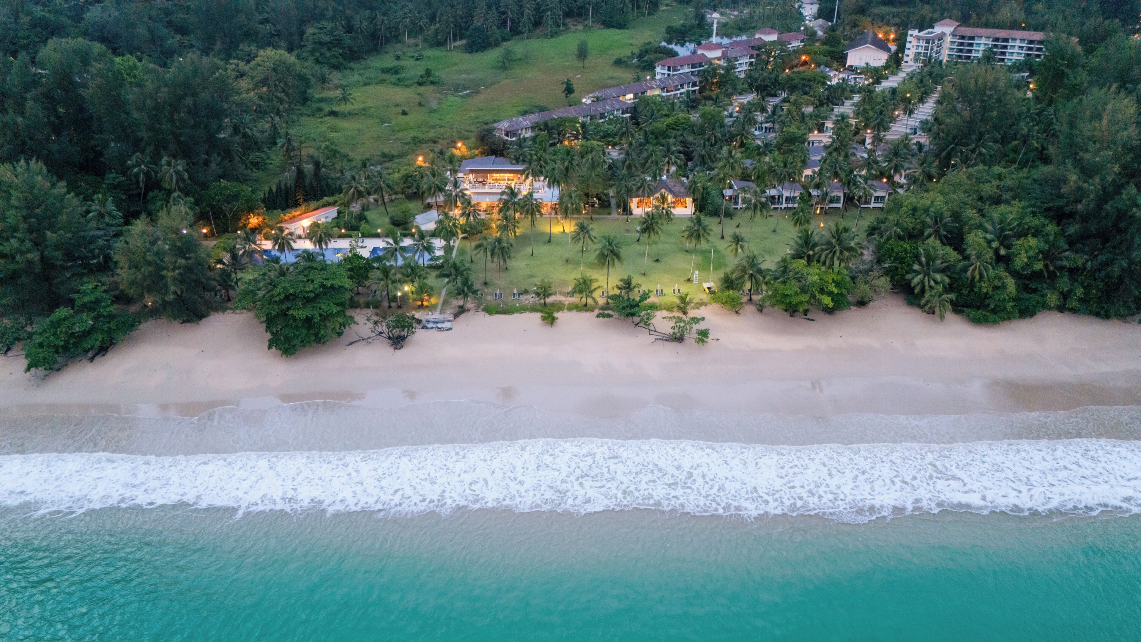 An aerial view of a coastal resort with beachfront properties, a sandy beach, and a backdrop of dense greenery.