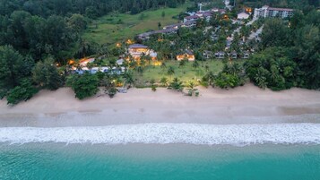 An aerial view of a coastal resort with beachfront properties, a sandy beach, and a backdrop of dense greenery.