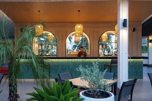 Zona de bar al aire libre con un bartender, techo de madera y vistas a palmeras a través de amplios ventanales.