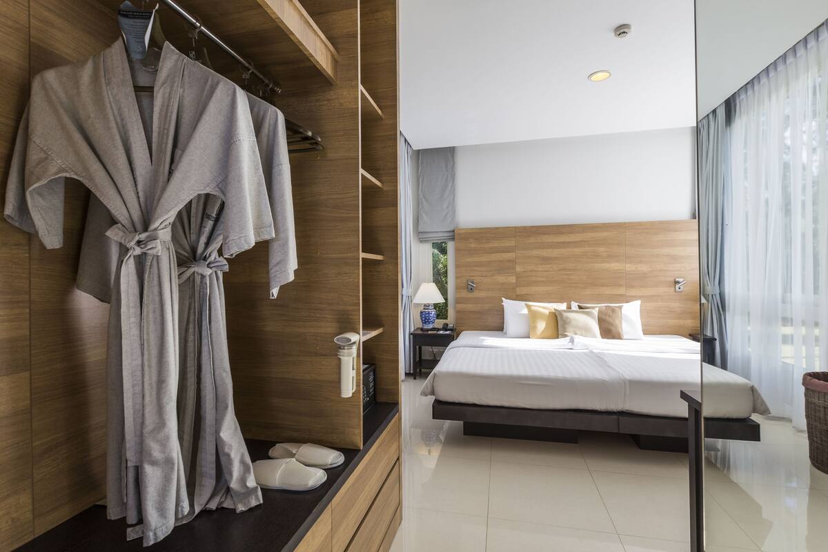 A modern hotel room with a wooden wardrobe, a bed with white bedding, a bedside table with a lamp, and a large window with curtains.