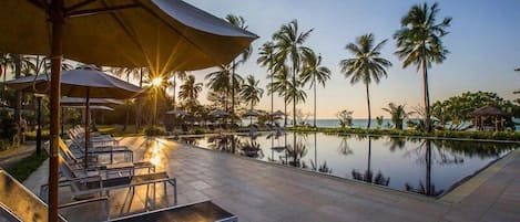 A poolside area with lounge chairs and umbrellas, palm trees, and a view of the water.