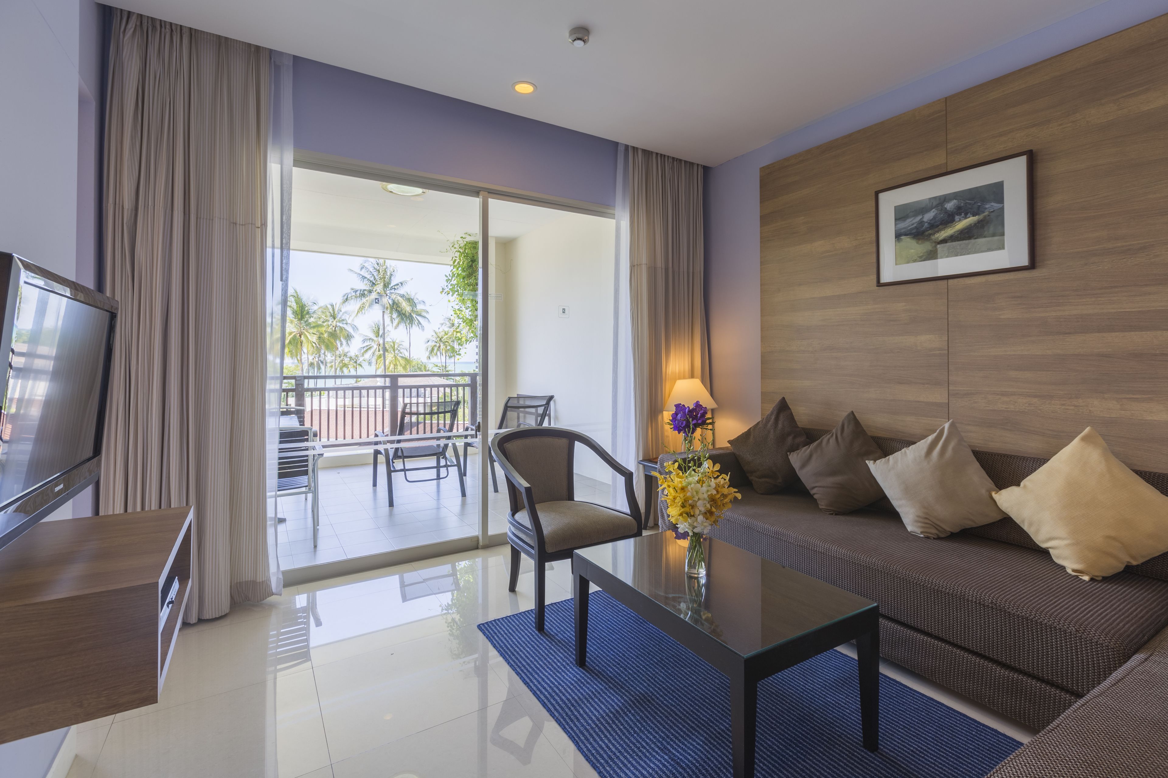 A modern hotel room with a sofa, armchair, and a TV on a wooden cabinet. A balcony with a view of palm trees is visible through the glass doors.