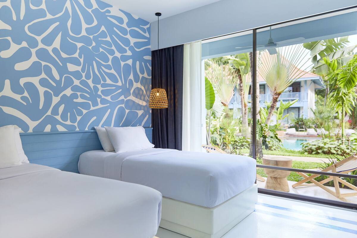 Ein Hotelzimmer mit zwei Betten, einem großen Fenster mit Blick auf einen Pool und viel Grün, sowie einer Wand mit Muster.