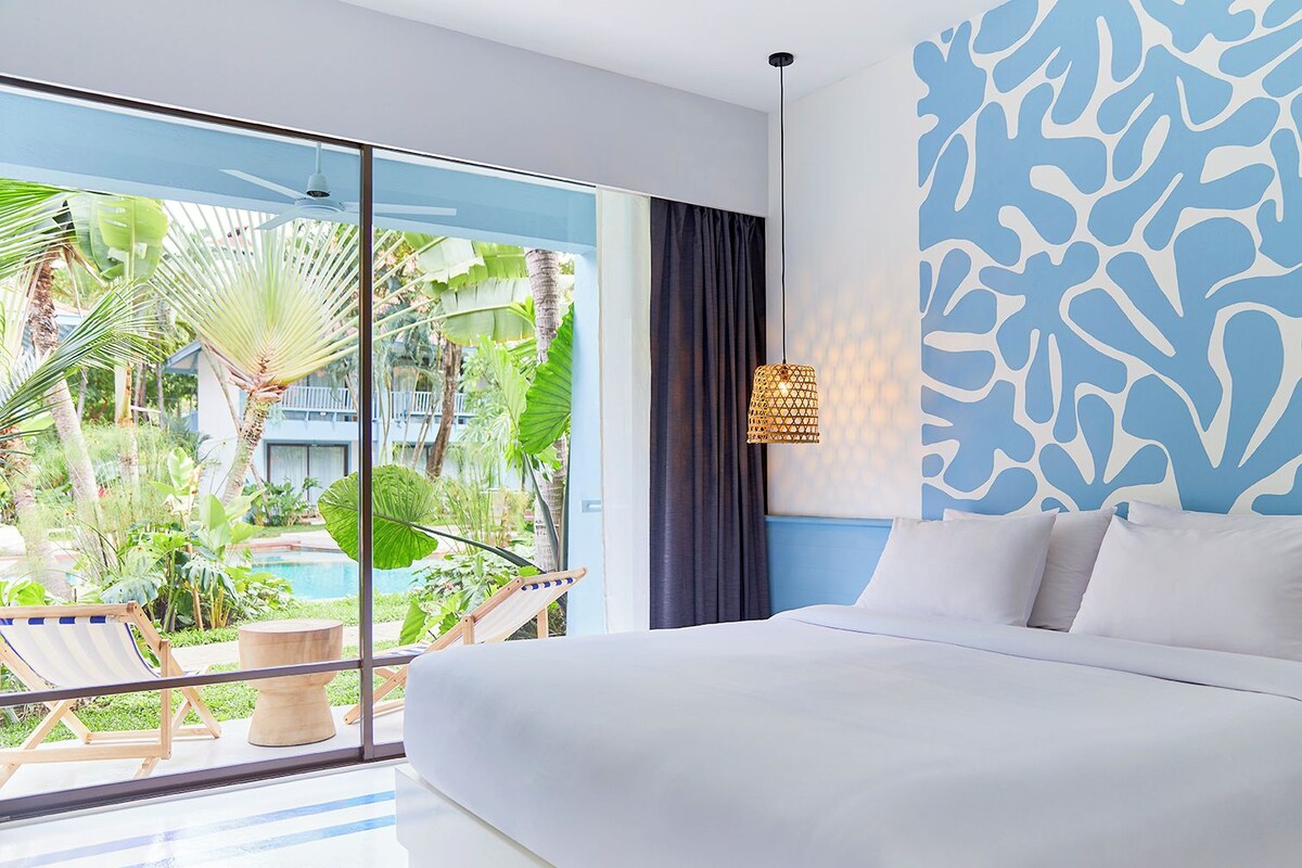 Une chambre d’hôtel moderne dotée d’un grand lit, offrant une vue sur une piscine et un jardin tropical, et présentant un décor original avec un mur bleu.