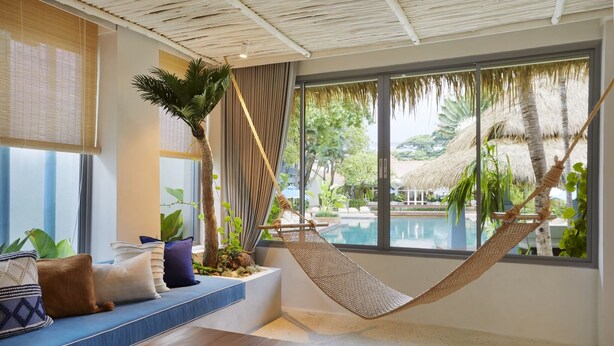 A room with a hammock, a seating area with cushions, a large window with a view of a pool and greenery. - The Peri Hotel Hua Hin (Hua Hin)