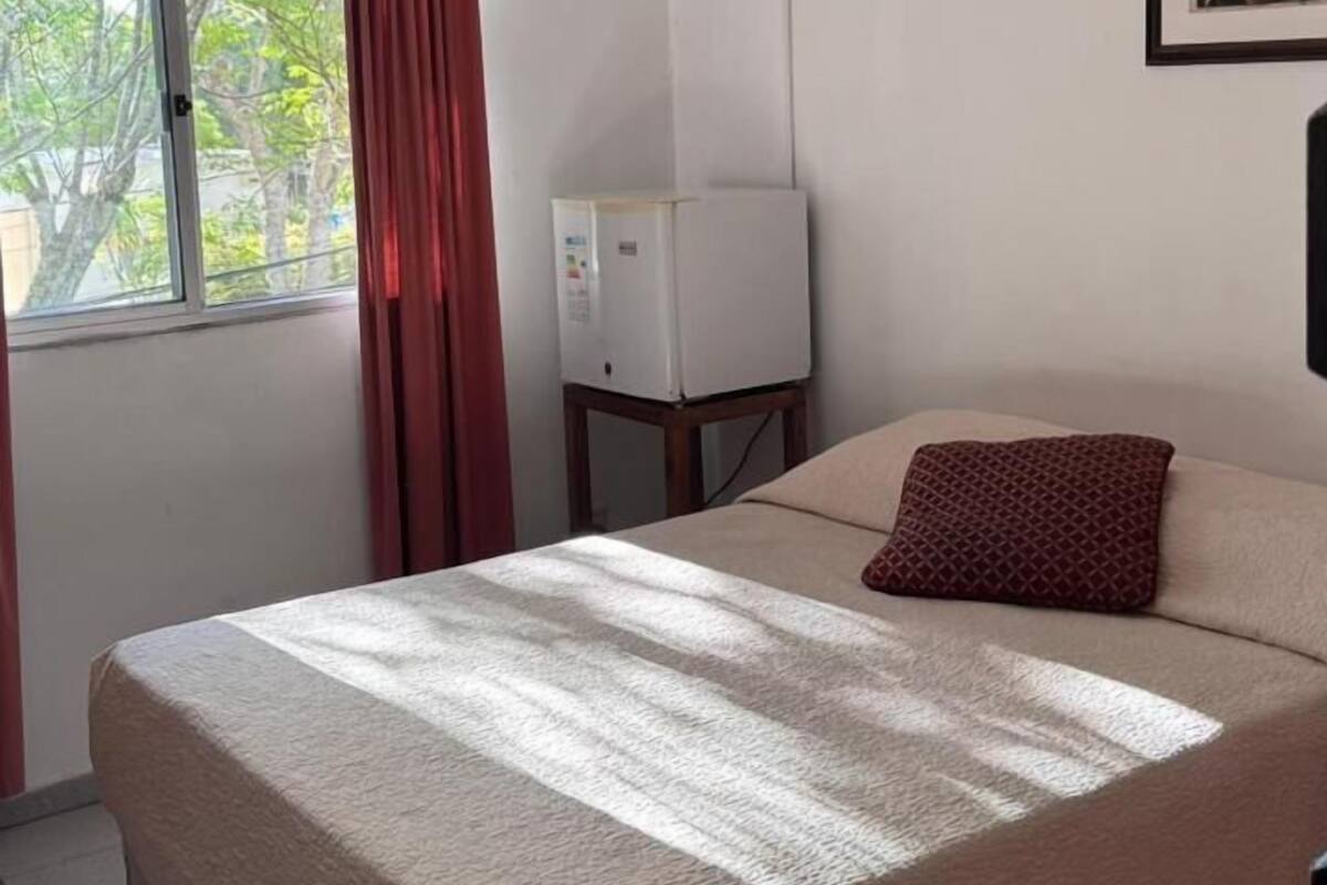 A bedroom with a bed, a window with curtains, a wall-mounted TV, and a ceiling light.