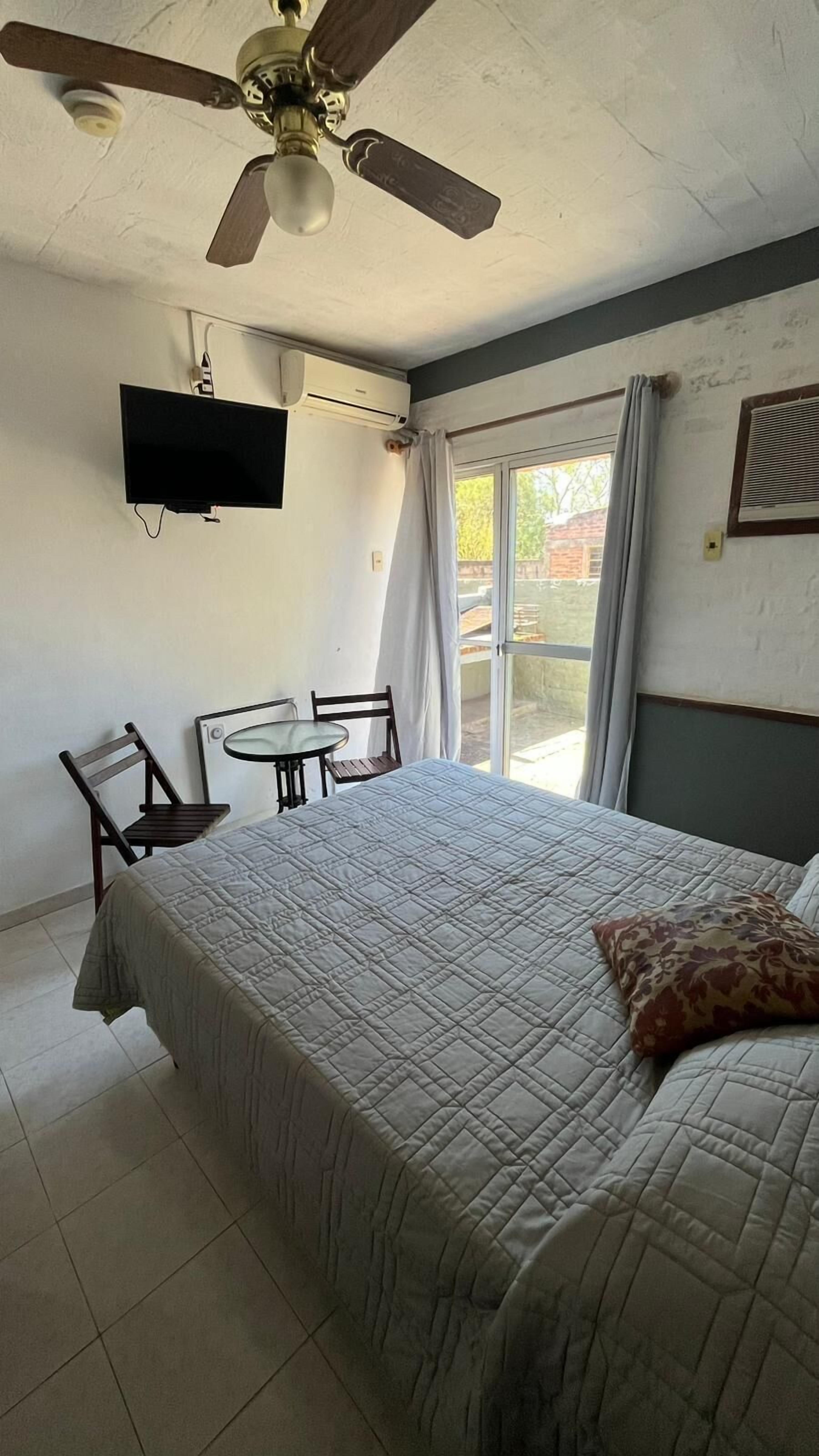 Un dormitorio con cama, ventilador de techo, televisor montado en la pared, una mesita con sillas y una ventana con cortinas.
