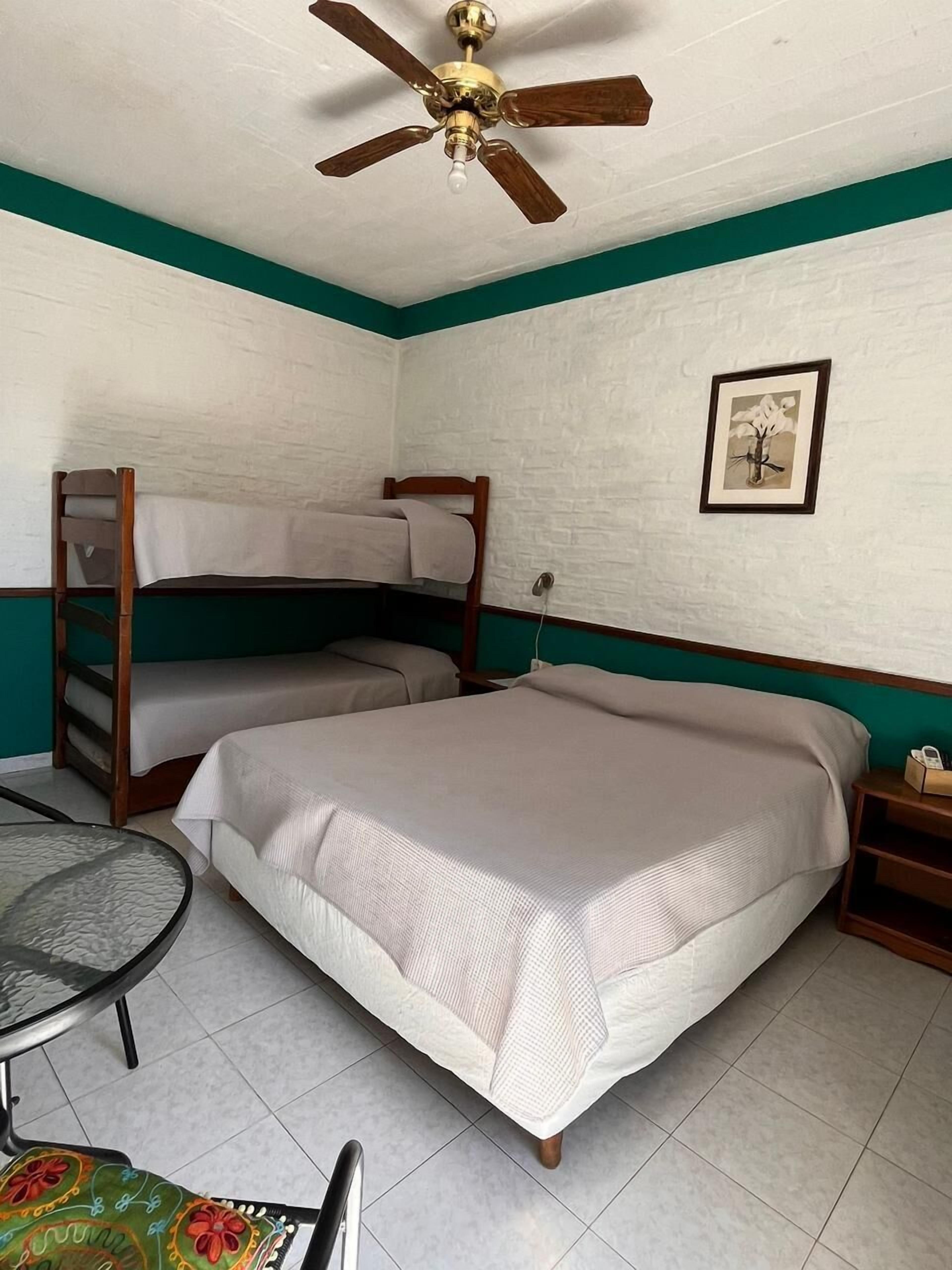 Una habitación con dos camas, un ventilador de techo, un cuadro en la pared y una pequeña mesa con una silla.