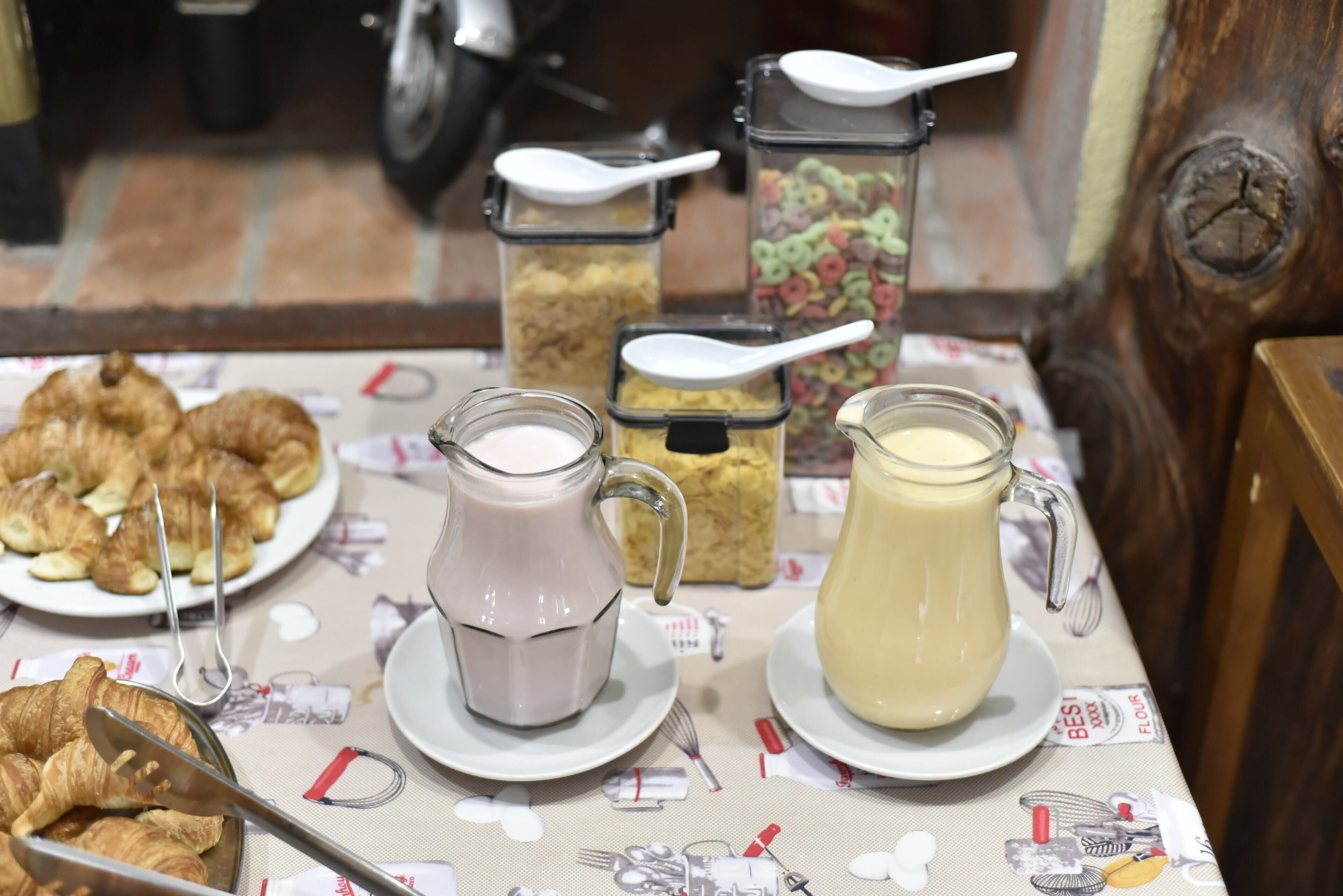 A breakfast table with croissants, two pitchers of milk, and cereal containers.