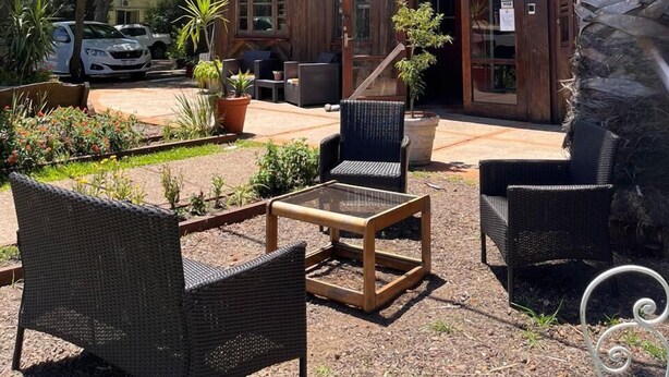 A two-story building with a wooden facade, outdoor seating area with wicker chairs and a table, and a palm tree to the right.