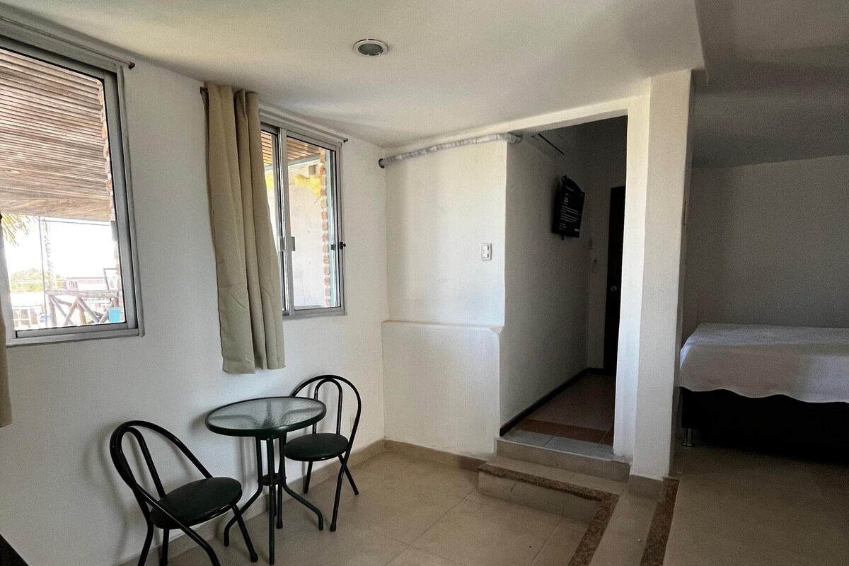 Een kamer met een bed, een klein tafeltje met twee stoelen, een raam met gordijnen en een trap naar een andere kamer.