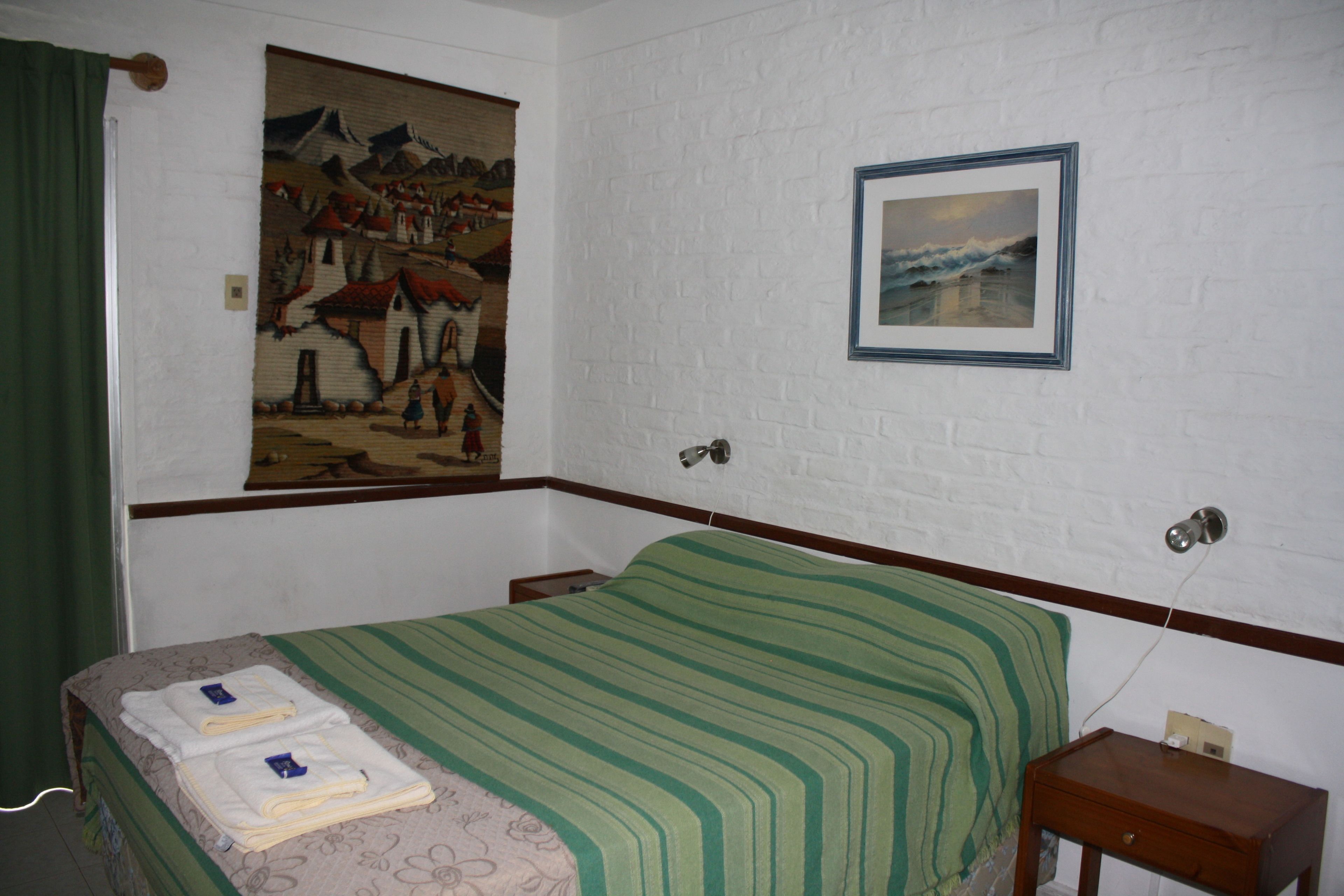 A bedroom with a bed, a painting of a village, a framed picture on the wall, and a small wooden nightstand.
