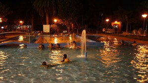 A nighttime swimming pool with people enjoying the water, surrounded by trees and street lamps.